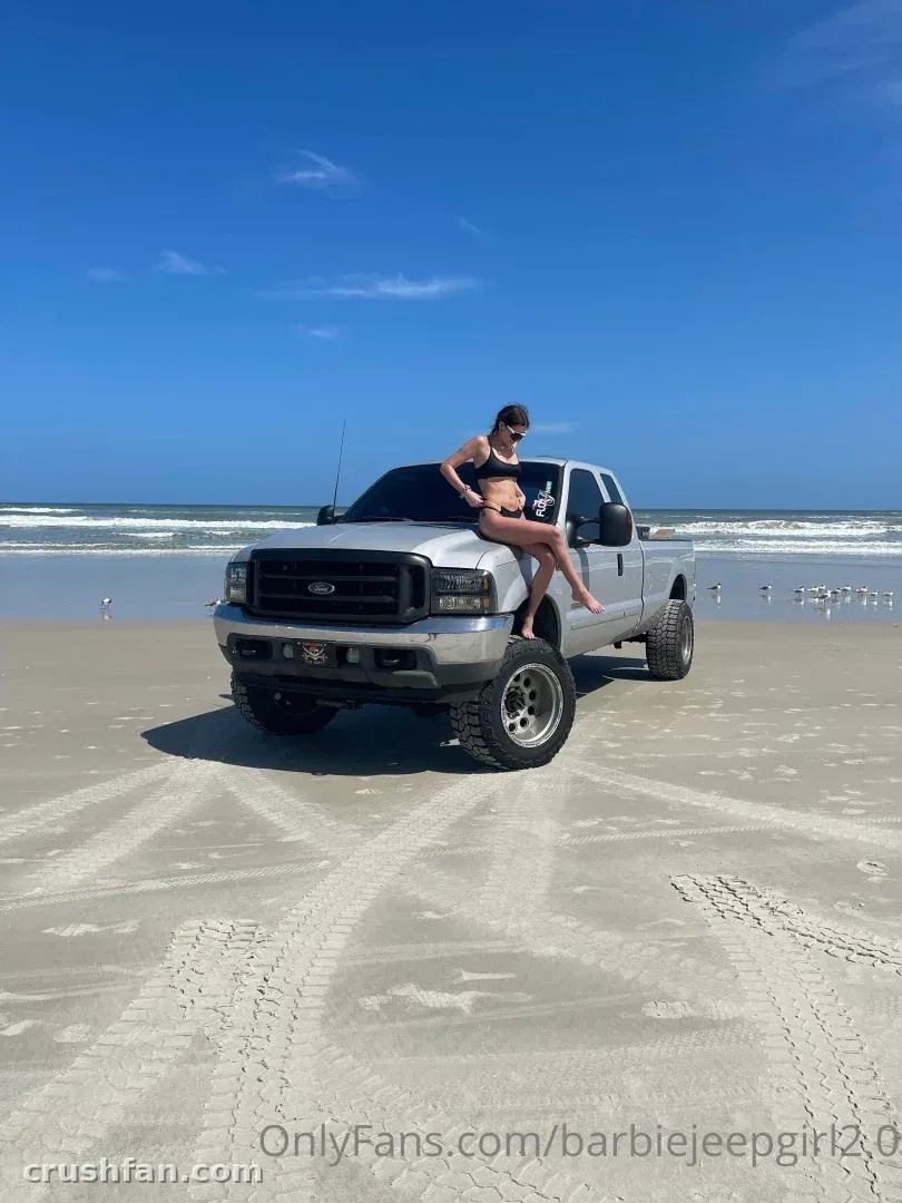 Woman in a dark swimsuit sitting on the hood of a silver truck on a sunny beach