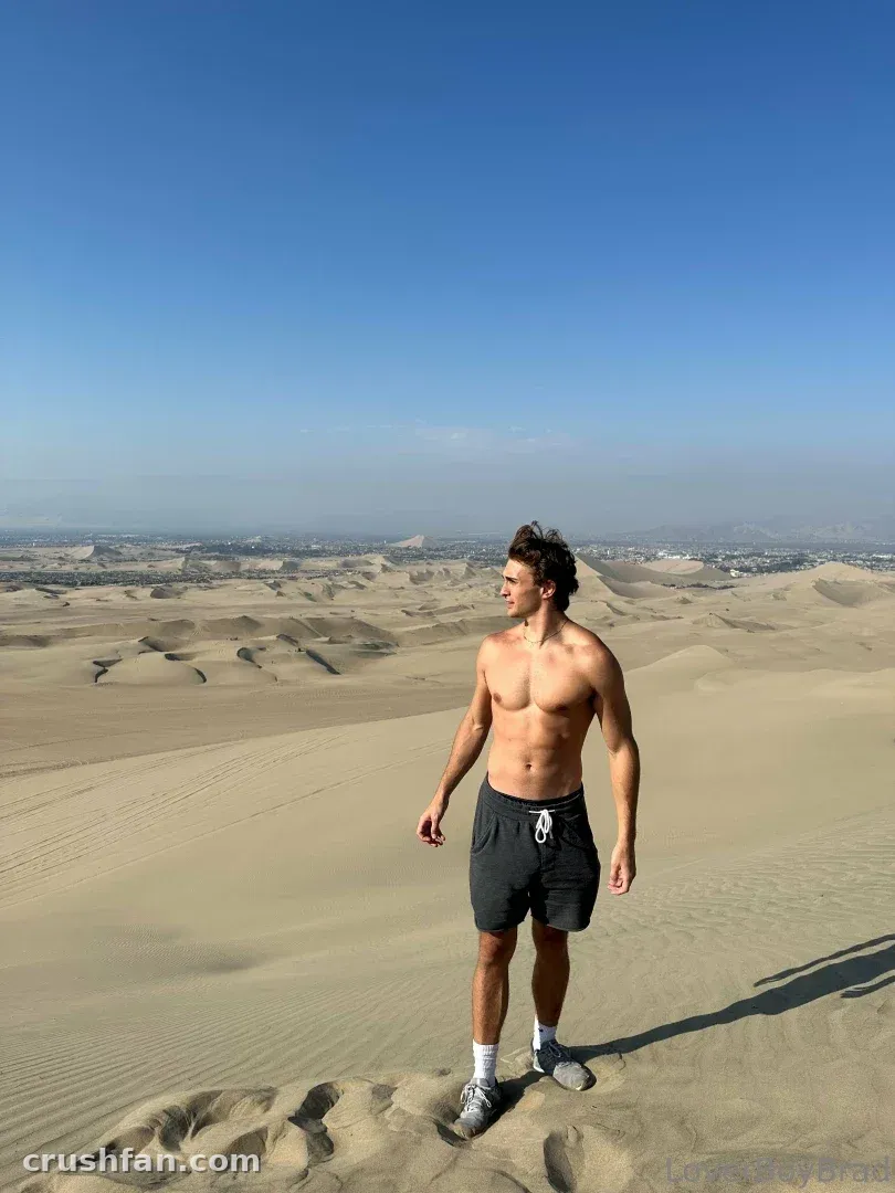 Leo shirtless in dark shorts standing on a desert sand dune under a blue sky