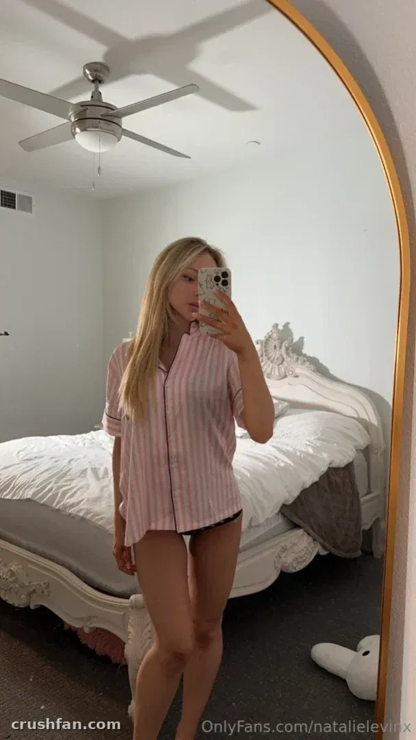 Natalie in a striped shirt and dark bottoms taking a mirror selfie in a bedroom.