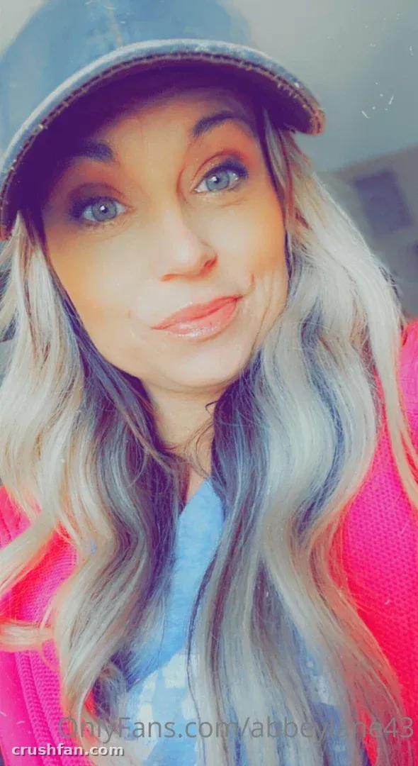 Selfie em close-up de Abbey Lane com boné escuro e cardigã rosa