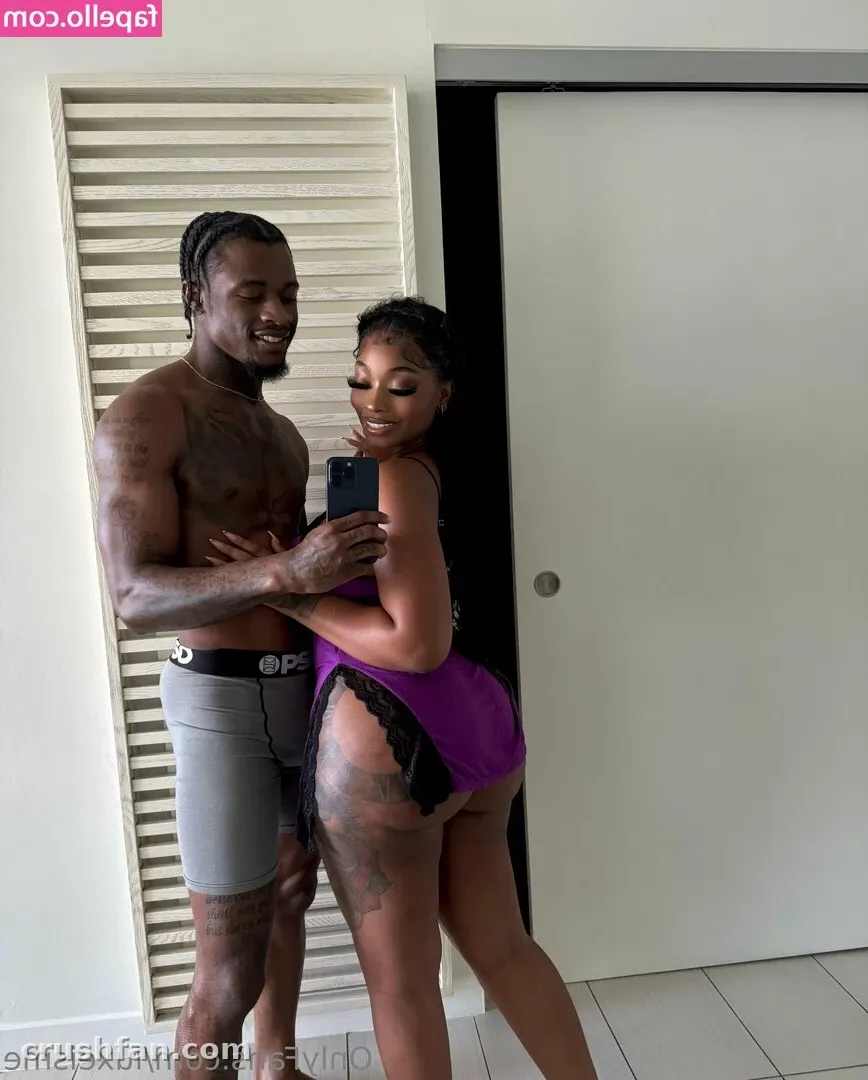Famousluxe, de vestido roxo com detalhe de renda, tirando uma selfie no espelho em ambiente interno ao lado de um homem sem camisa.