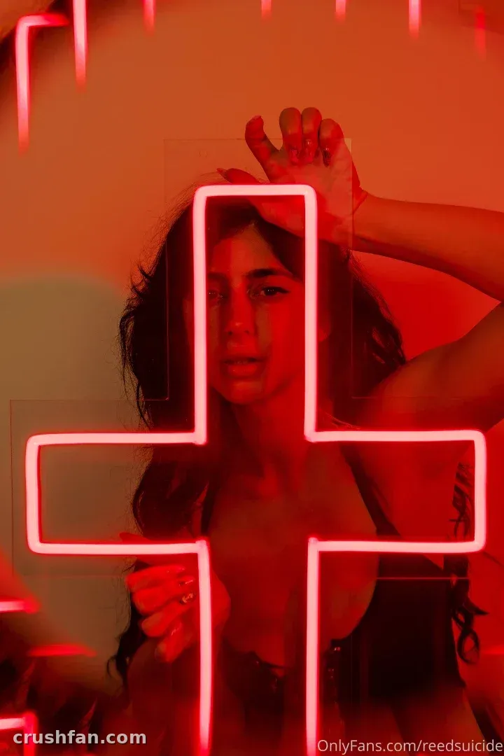 Woman with dark hair behind a glowing red neon cross in a dim room