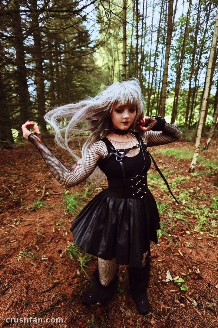 Creator in a black lace-up dress and fishnets posing among tall forest trees