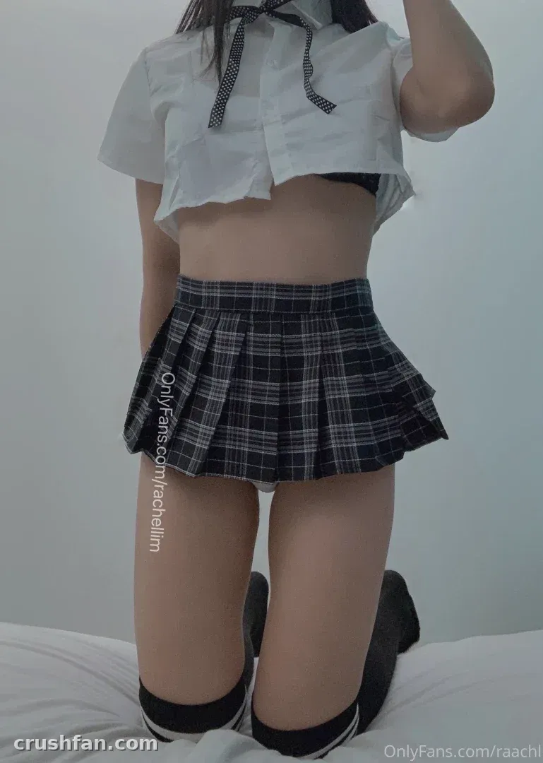 Rachel kneeling on a bed in a white cropped shirt, plaid pleated skirt and dark knee socks.
