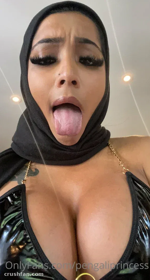 Yasmina Khan avec un foulard noir sur la tête et un haut brillant, tirant la langue de façon espiègle dans un selfie en gros plan.