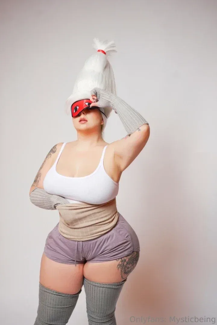 MysticBeing in white top, purple shorts, red mask and grey sleeves against a plain wall