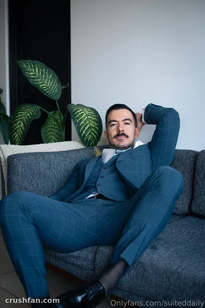Suited Daily in a blue three-piece suit reclining on a grey sofa beside a green plant