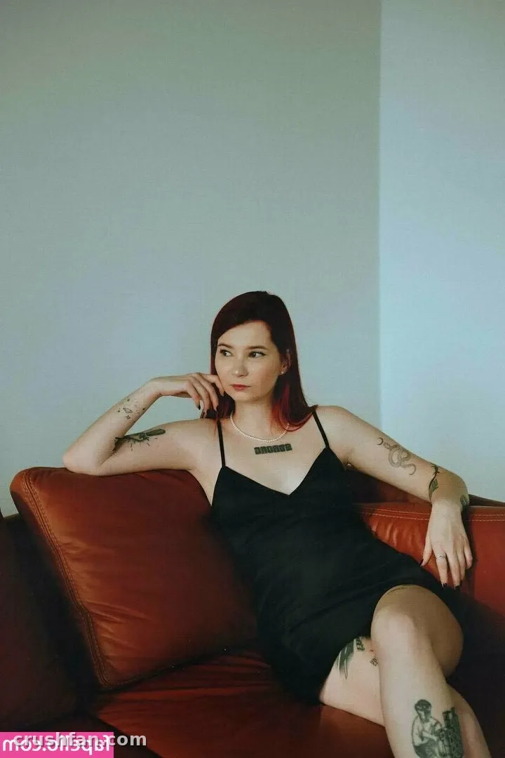 Anicka com um vestido slip preto reclinada em um sofá de couro marrom-avermelhado contra uma parede clara