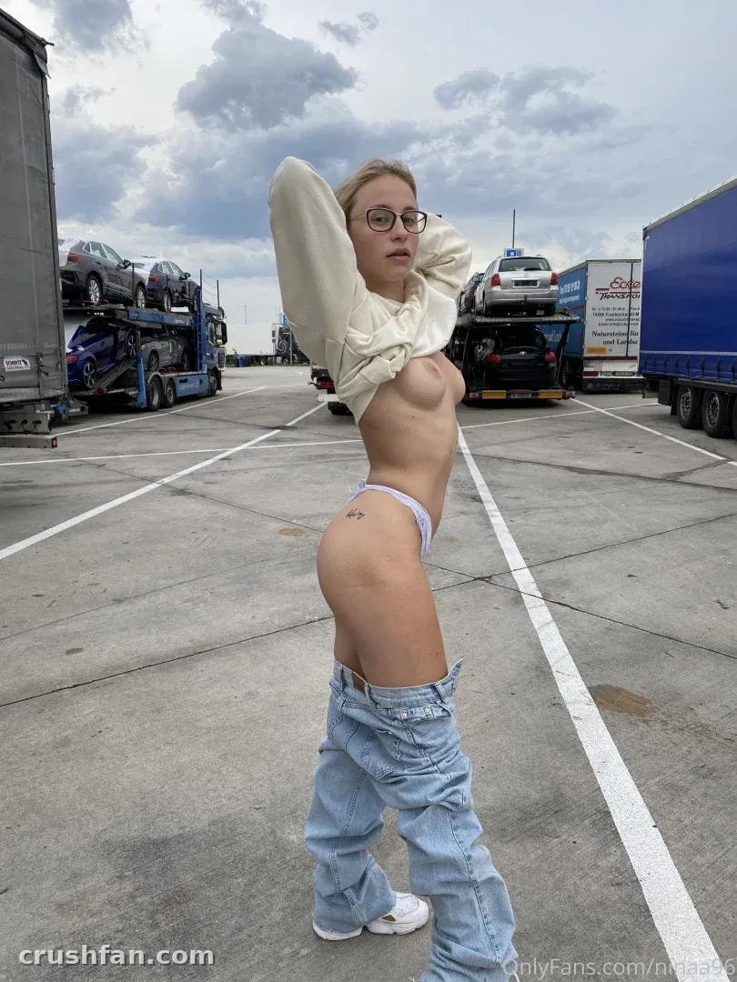 Nina Joy de pie en un aparcamiento pavimentado de vehículos bajo un cielo nublado, con vaqueros holgados y una prenda superior de color crema.