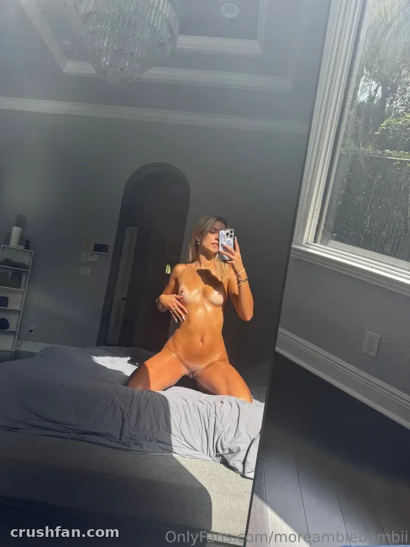 Ambie Bambie prenant un selfie miroir agenouillée sur un lit dans une pièce baignée de soleil.