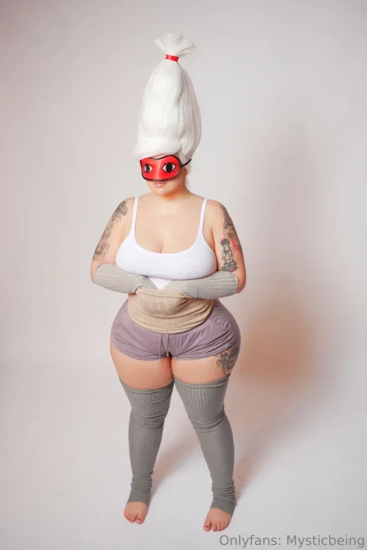 MysticBeing in einem weißen Top, grauer Shorts und oberschenkelhohen Socken vor einem schlichten Hintergrund