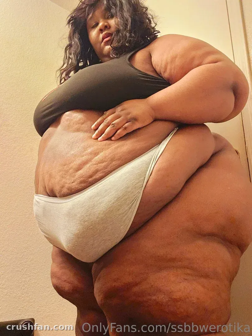 Erotika in a dark tank top and light briefs, photographed from a low angle indoors.