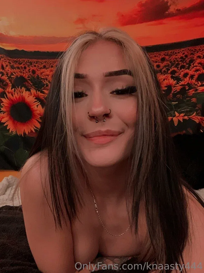 A criadora KNA$TY sorrindo em frente a uma tapeçaria de girassóis com tons quentes de céu laranja.