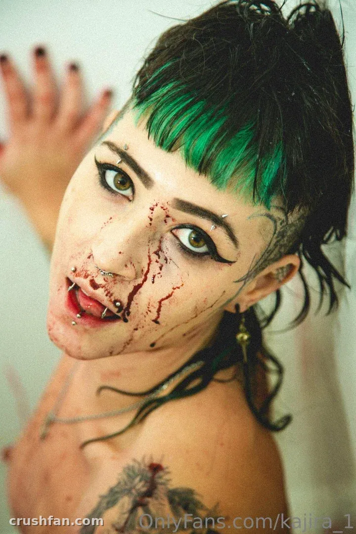 Kajira com franja verde, delineador escuro e maquiagem de sangue falso, mão levantada perto do rosto