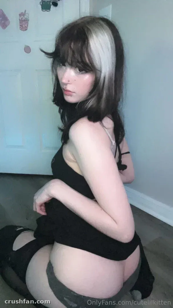 Aba kneeling indoors in a black top and stockings, looking back over her shoulder.