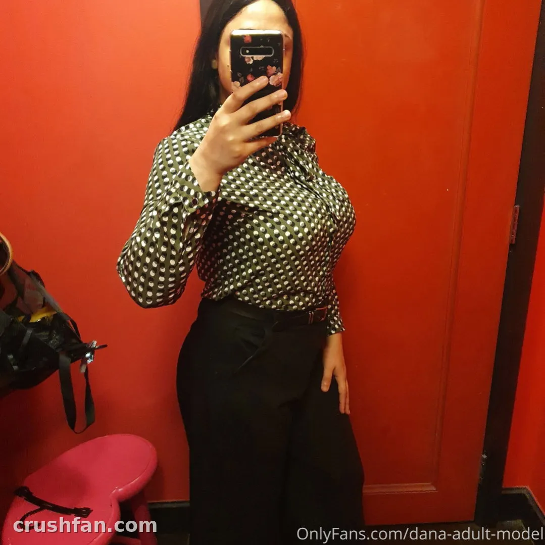 Creator in a patterned blouse and dark trousers taking a mirror selfie against a red wall