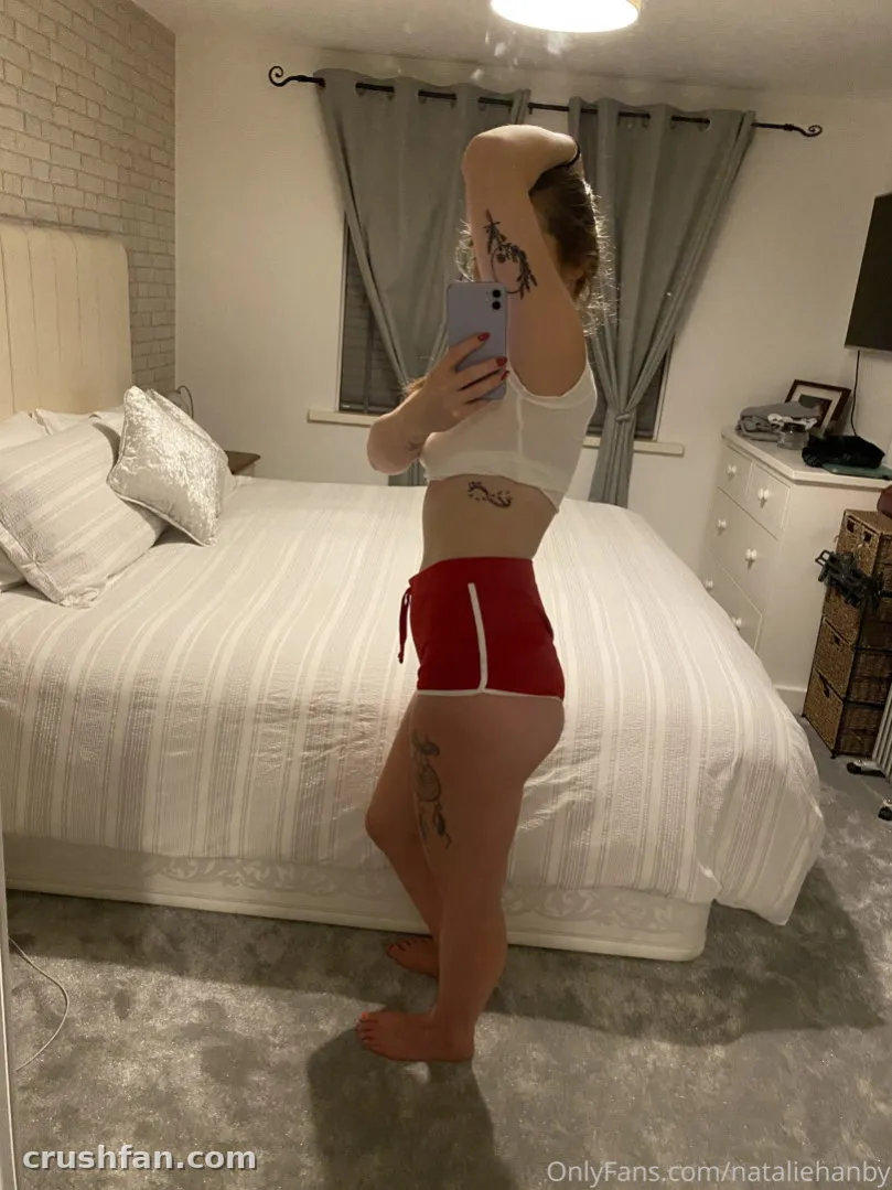 Natalie Hanby in a white top and red shorts taking a mirror selfie in a bedroom.