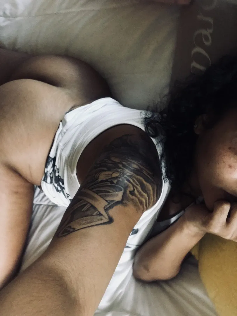 Bella Cream lying on white bedding with a large tattoo on her arm