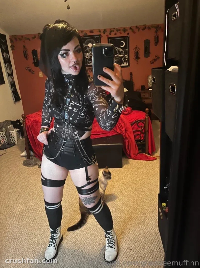 Ghostiee in a black graphic top and skirt posing in a mirror inside a bedroom with orange walls.