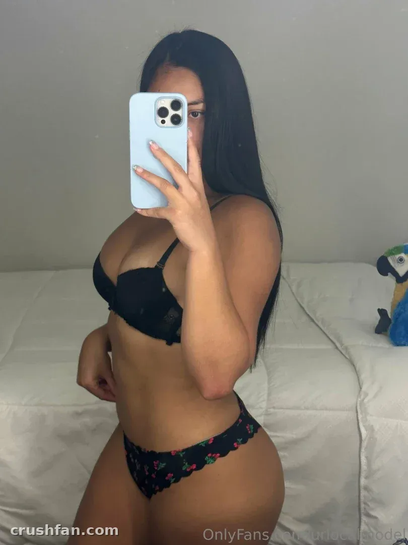 Creator in black lingerie taking a mirror selfie in a bright bedroom with white bedding.