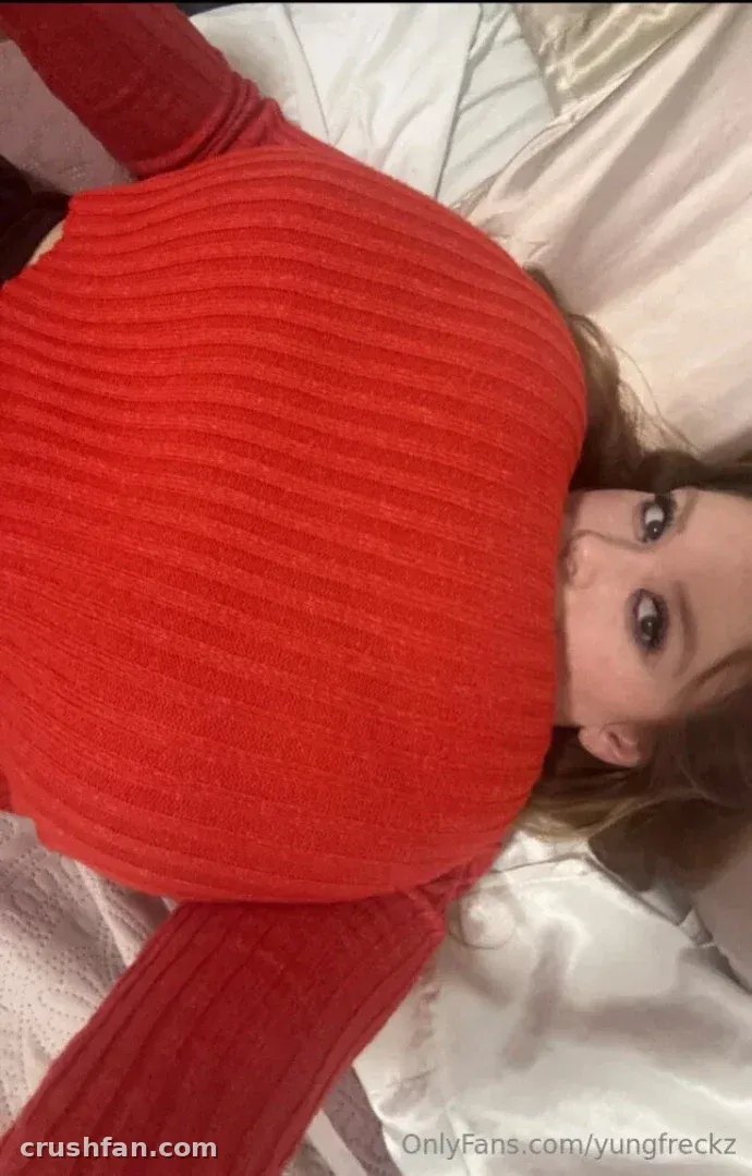 Missy con un top acanalado rojo recostada sobre ropa de cama blanca, mirando hacia la cámara