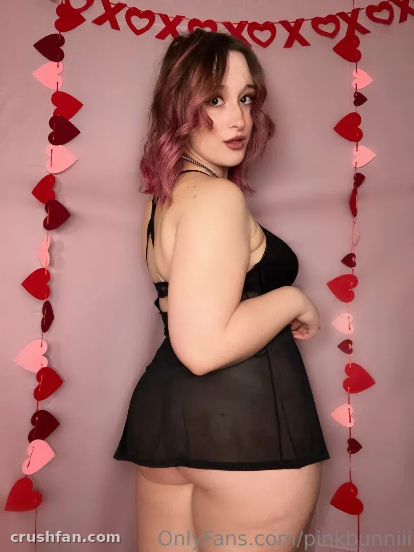 AryiaFawn en lingerie noire devant un fond rose avec des guirlandes de cœurs
