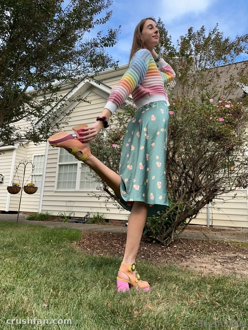 Ellie dans un pull rayé arc-en-ciel et une jupe florale bleu canard en équilibre sur une jambe dehors.