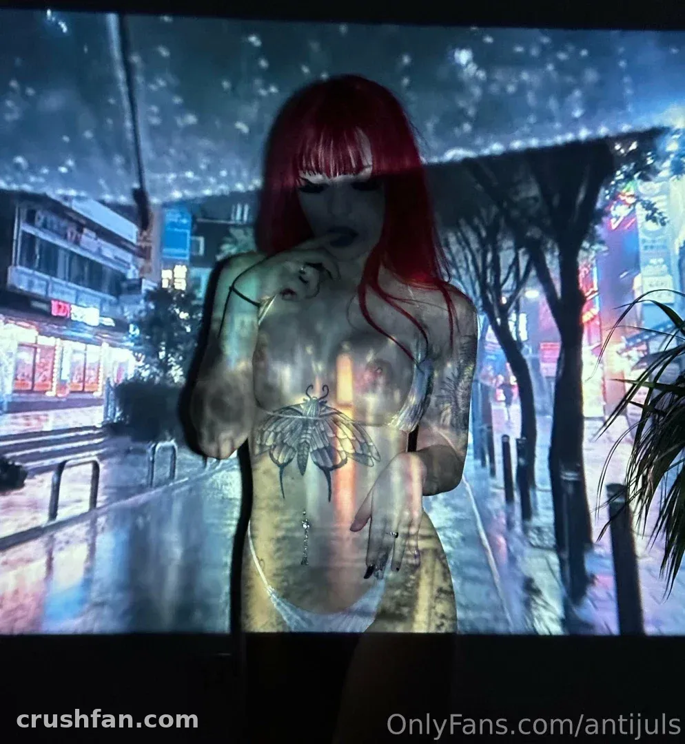 Juls com cabelo vermelho posando diante de uma projeção de rua urbana de neon